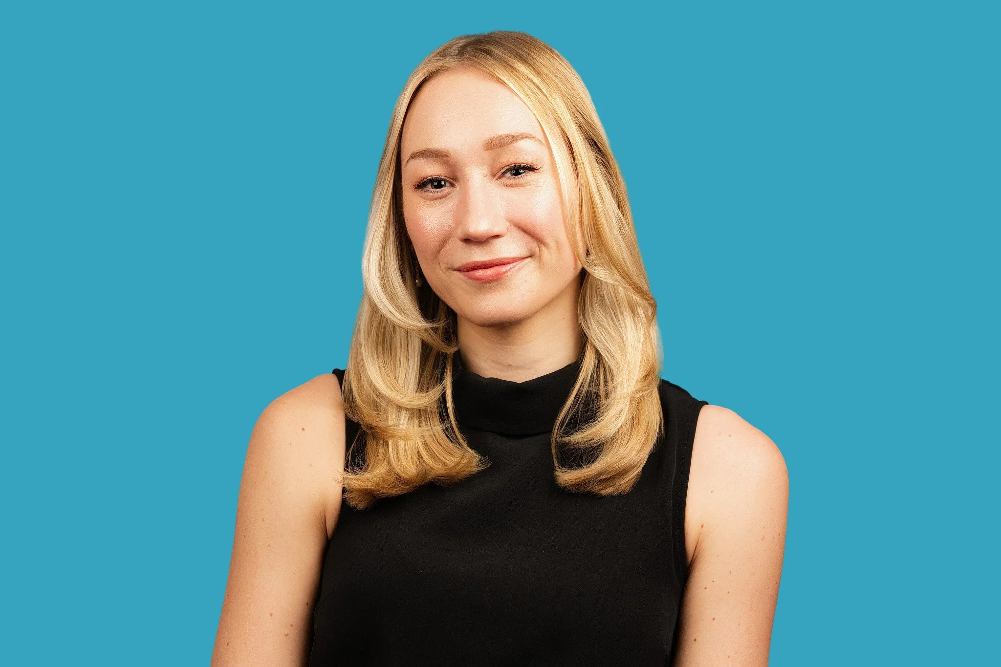 A woman with long blonde hair is wearing a black sleeveless turtleneck top, smiling softly, posed against a bright teal background.