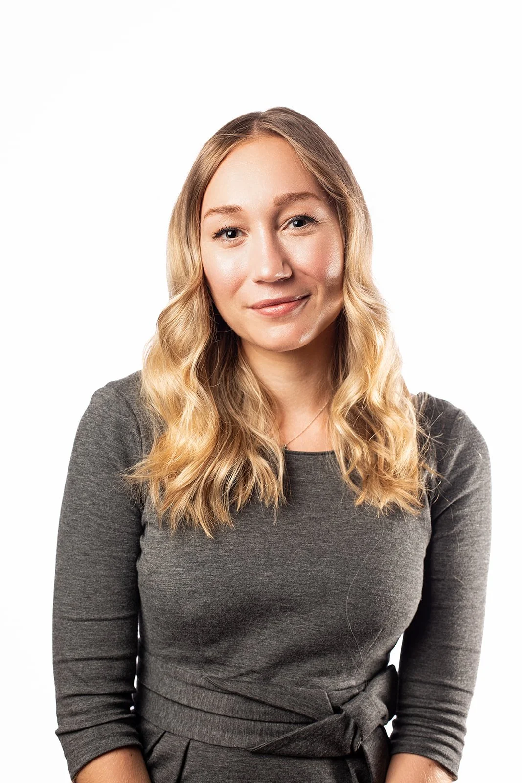 A young woman with blonde hair, smiling, wearing a dark gray dress, against a plain white background.