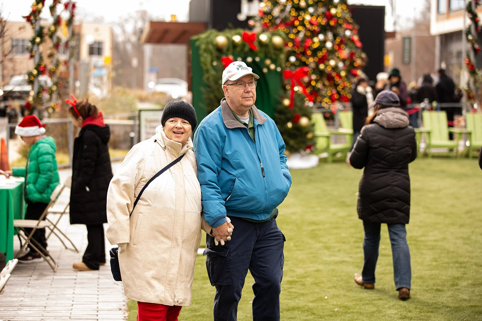 A happy couple holding hands and smiling at a Christmas outdoor event with a decorated Christmas tree in the background, surrounded by people wearing winter clothing.