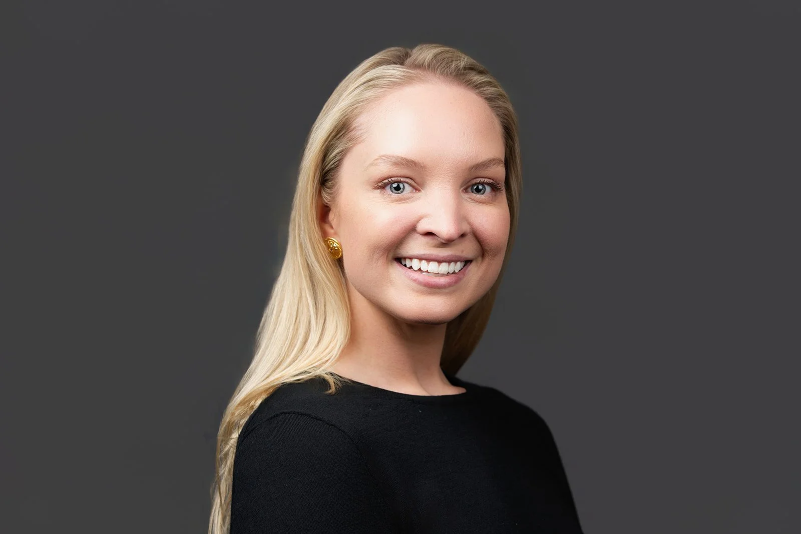 A young woman with long blonde hair, blue eyes, and a bright smile wearing gold earrings and a black top, against a dark grey background.