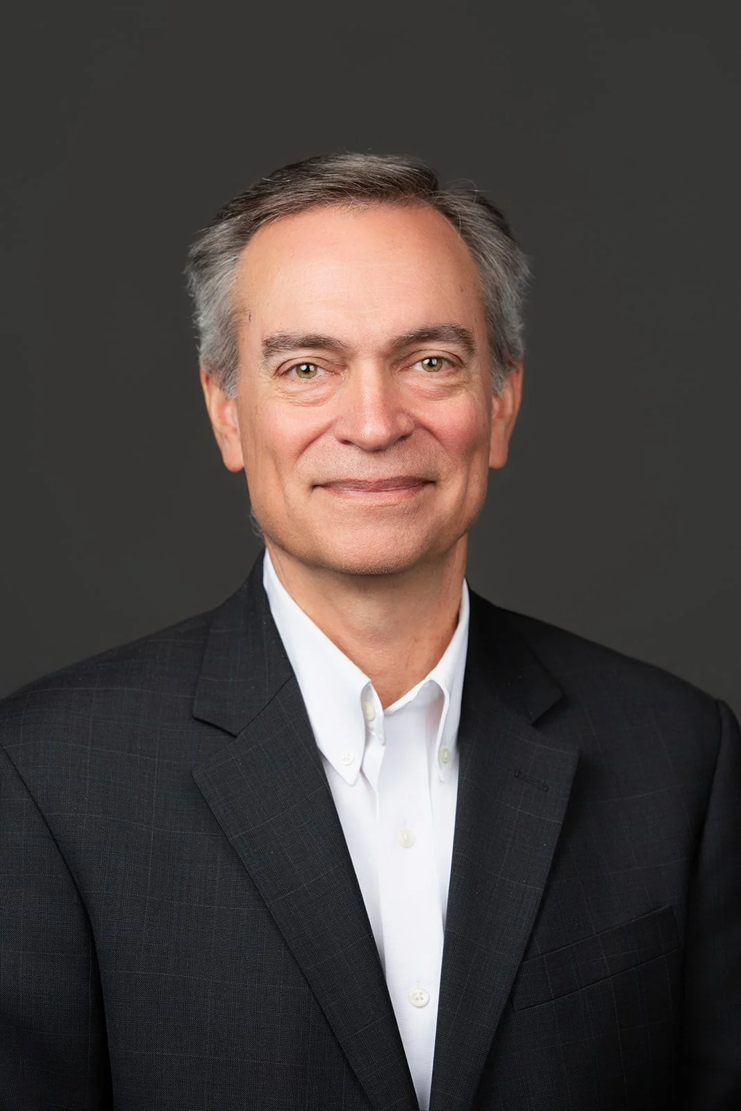 A professional portrait of a middle-aged man wearing a black suit jacket and a white dress shirt, against a dark gray background.