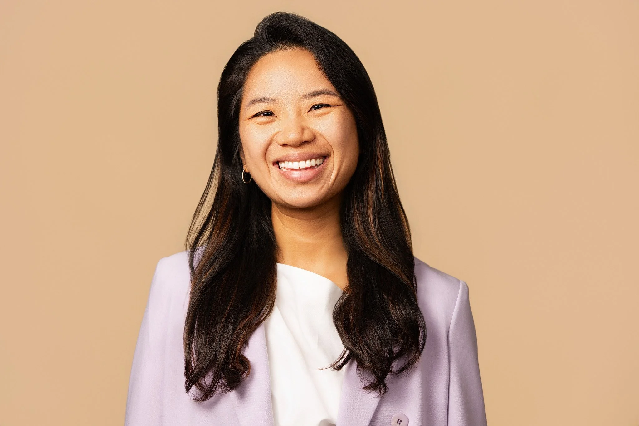 A smiling woman with long dark hair, wearing a light purple blazer and white top, standing against a beige background.