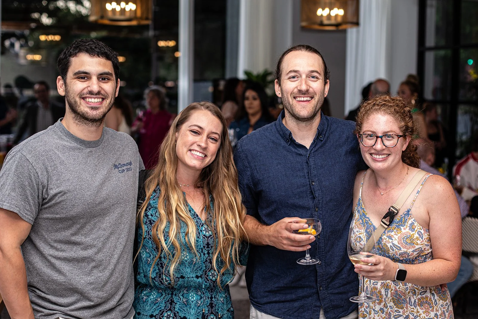 Four friends smile warmly at a social gathering, their closeness and relaxed expressions radiating joy and camaraderie. Two hold wine glasses, suggesting a celebratory atmosphere. The group stands out against a softly lit, blurred background that hin