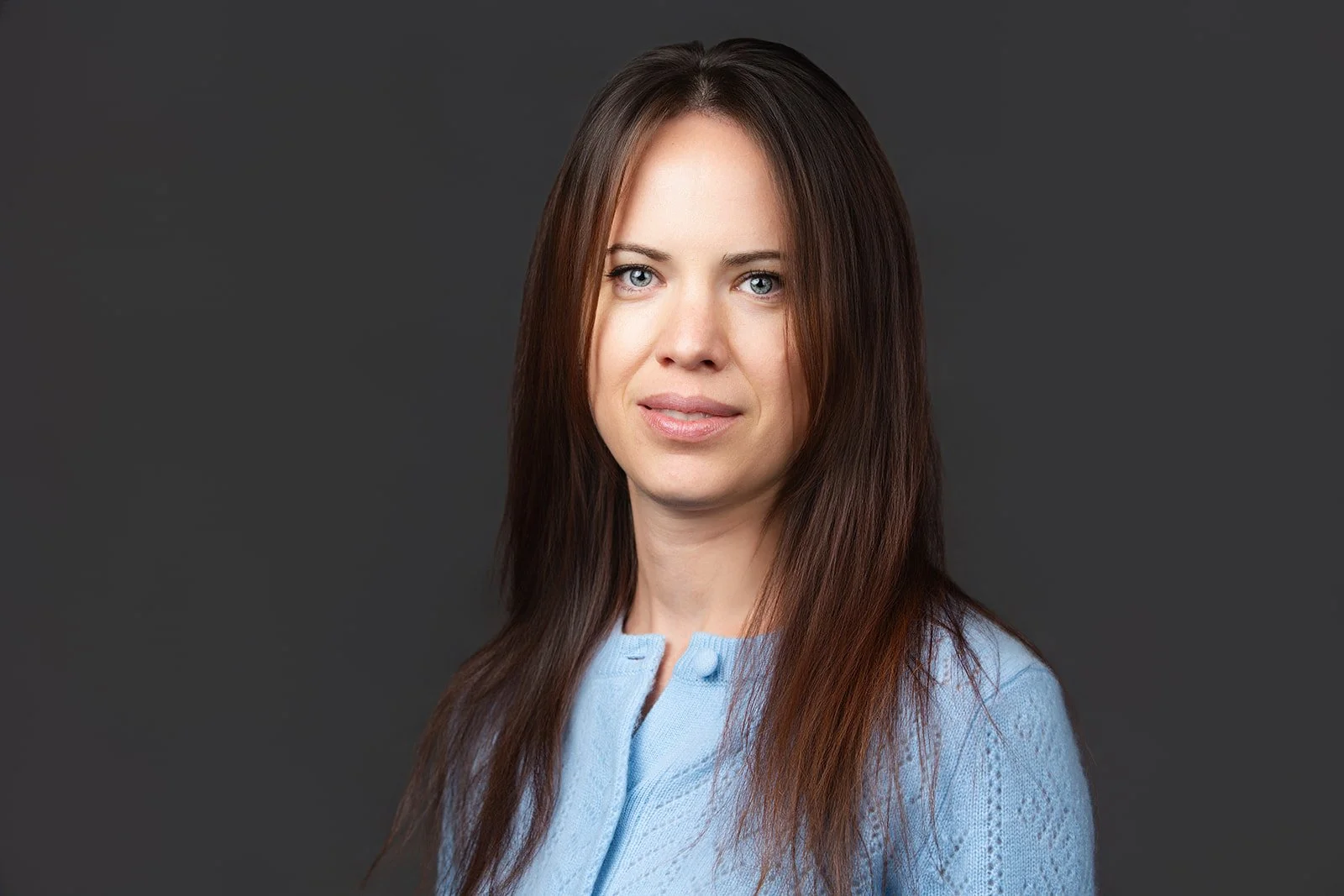 A woman with long brown hair wearing a light blue top in front of a dark gray background.