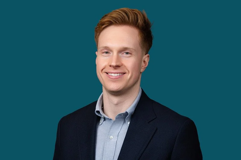 A professional headshot of a smiling man with red hair wearing a dark navy blazer and light blue dress shirt, photographed against a deep teal background.