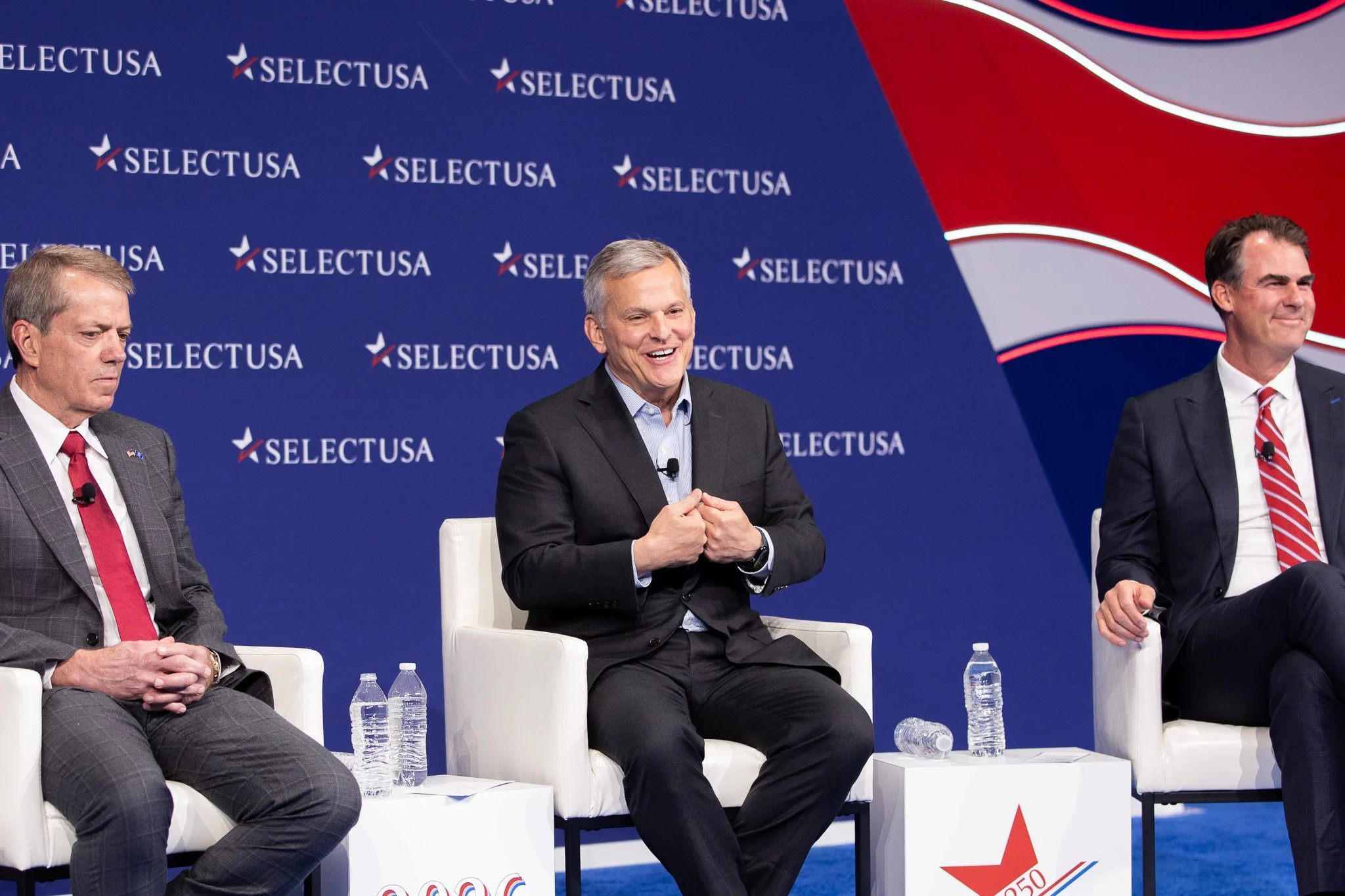Three executives participate in a panel discussion at the SelectUSA Investment Summit, sharing perspectives on business investment and economic development. Seated on a professionally designed stage with SelectUSA branding, the speakers engage in a l