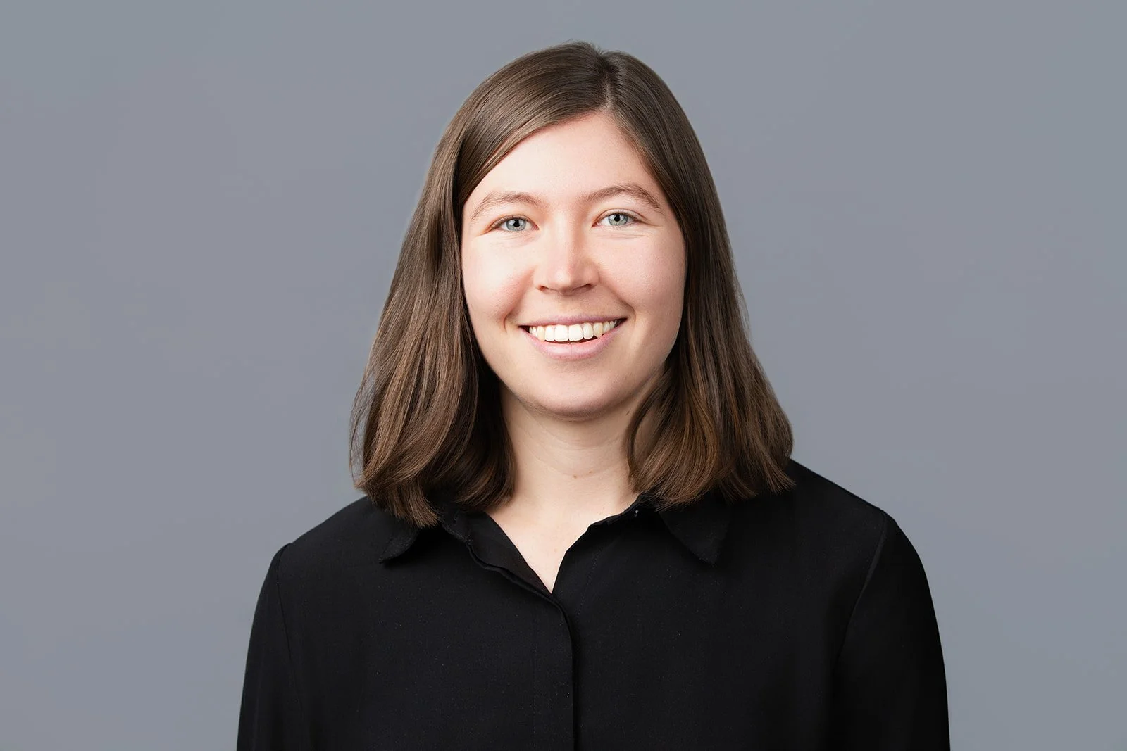 Woman with medium-length brown hair wearing a black button-down shirt, standing in front of a solid grey backdrop.