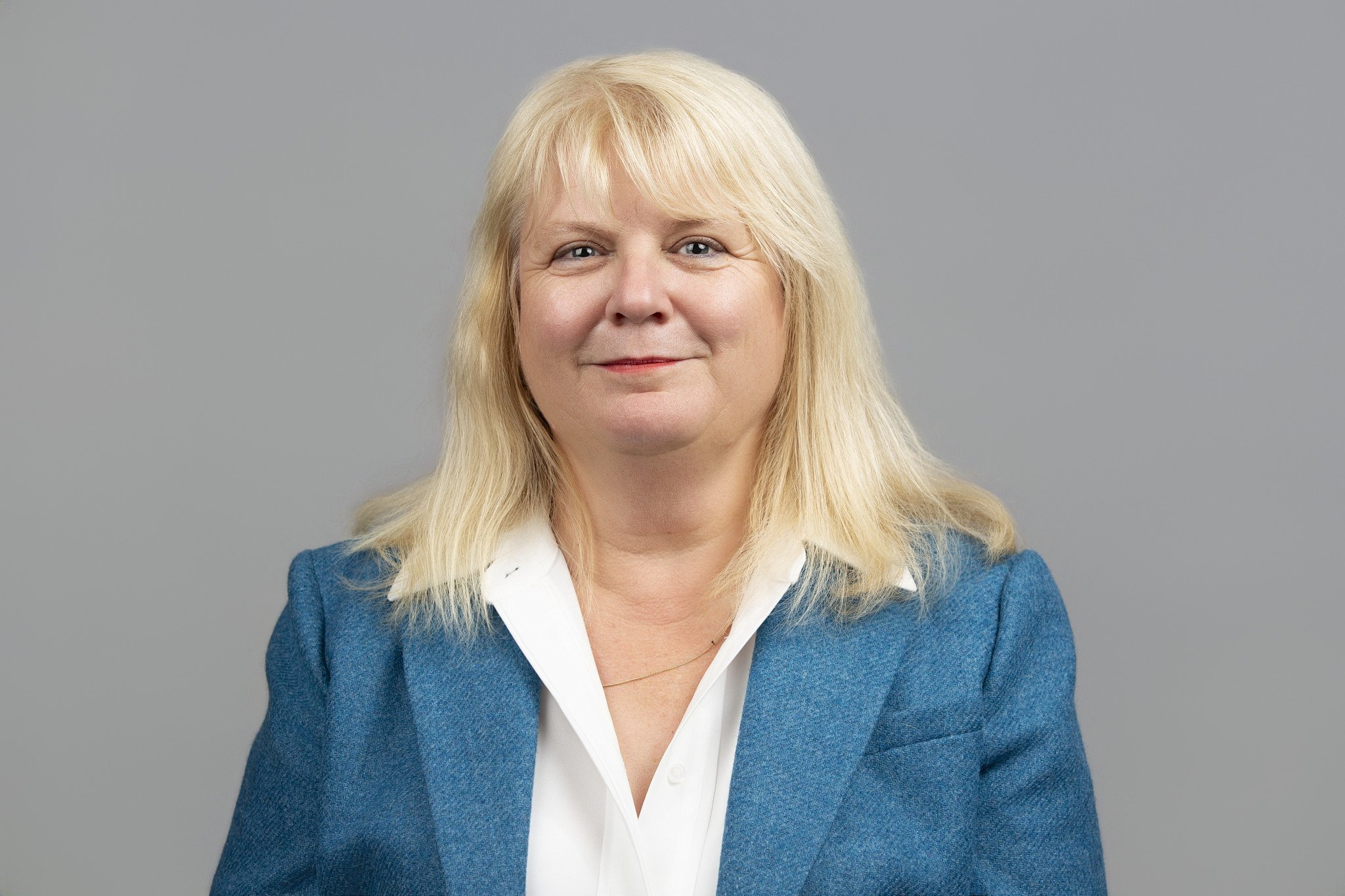 Portrait of a middle-aged woman with blonde hair, wearing a blue blazer and white collared shirt, smiling against a plain gray background.