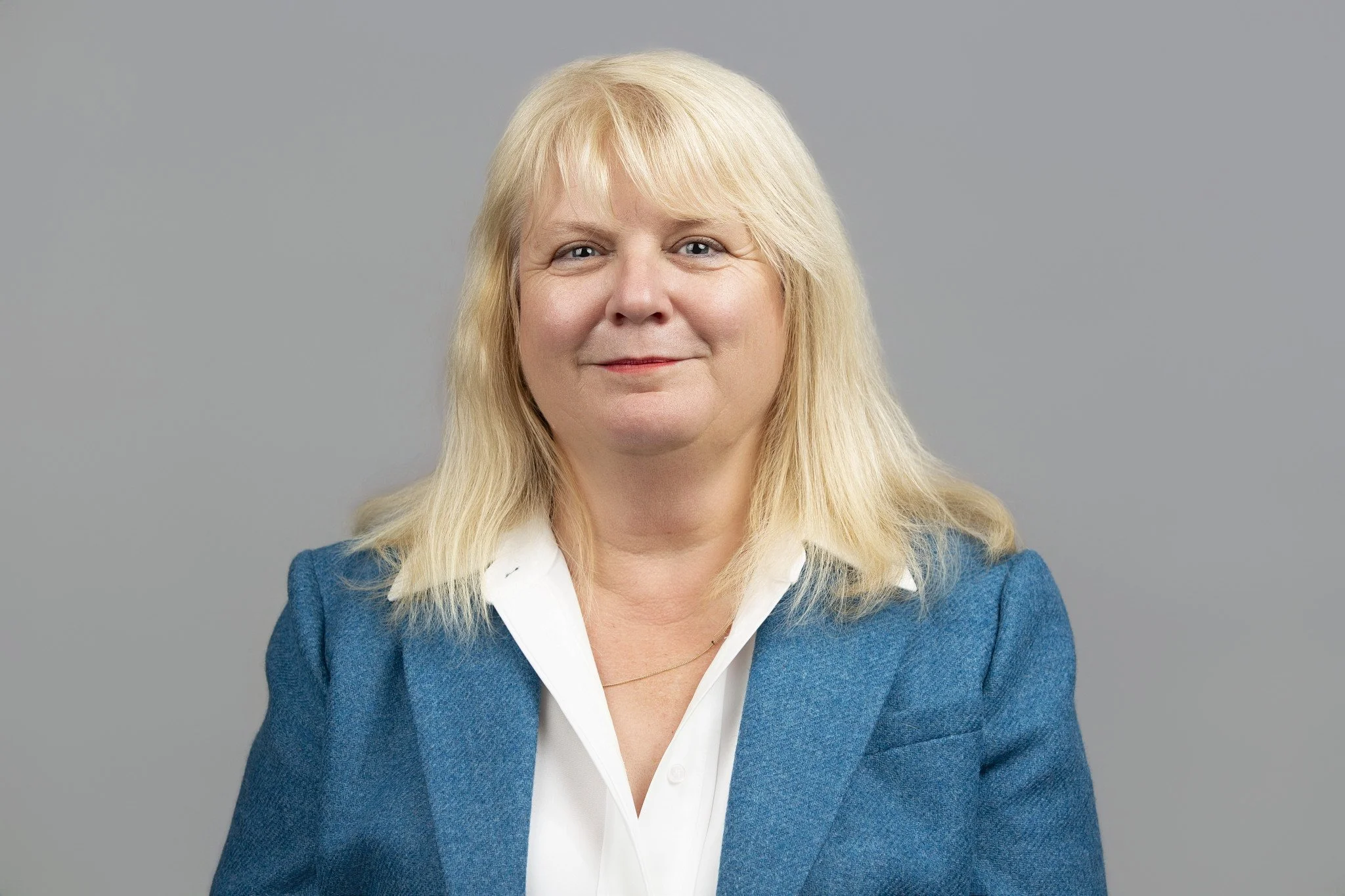 Middle-aged woman with blonde hair wearing a white collared shirt and a turquoise blazer, standing in front of a grey backdrop.