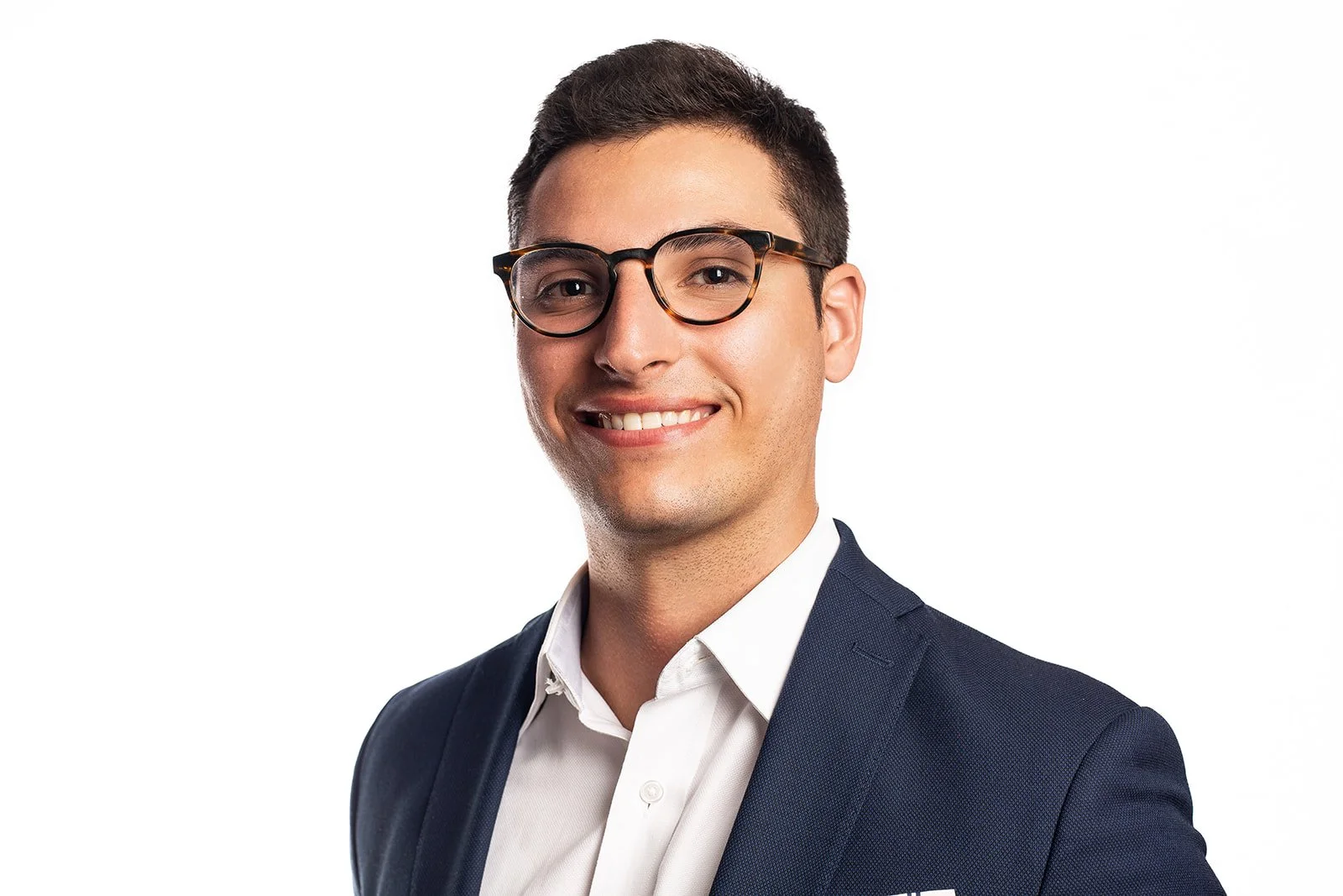 Portrait of a young man with dark hair, glasses, in a navy suit and white shirt, smiling against a white background.