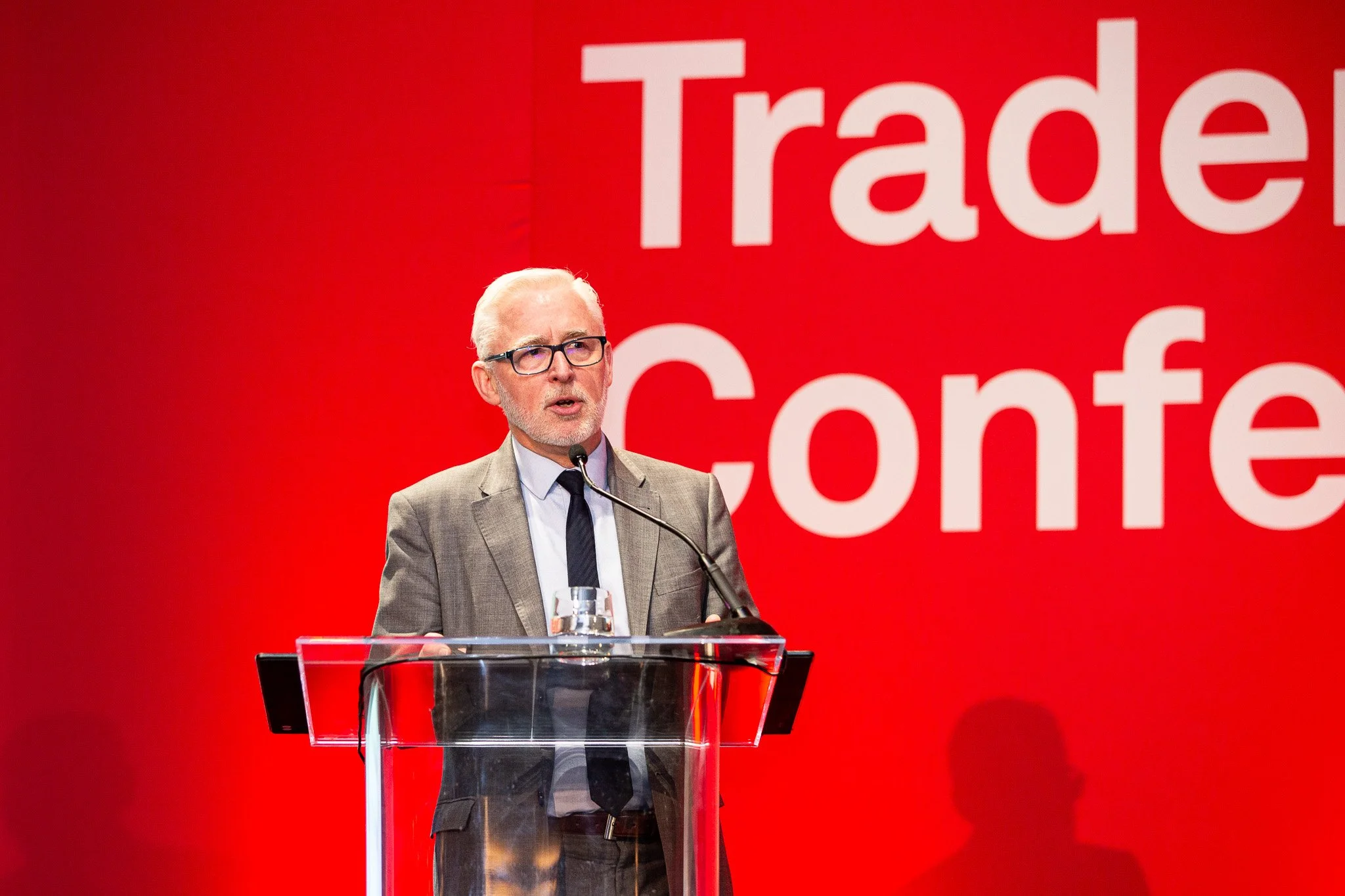 A keynote speaker delivers remarks from a podium during a professional trade conference, addressing an audience against a bold red event backdrop. The image captures a moment of public speaking and thought leadership, highlighting the energy, experti