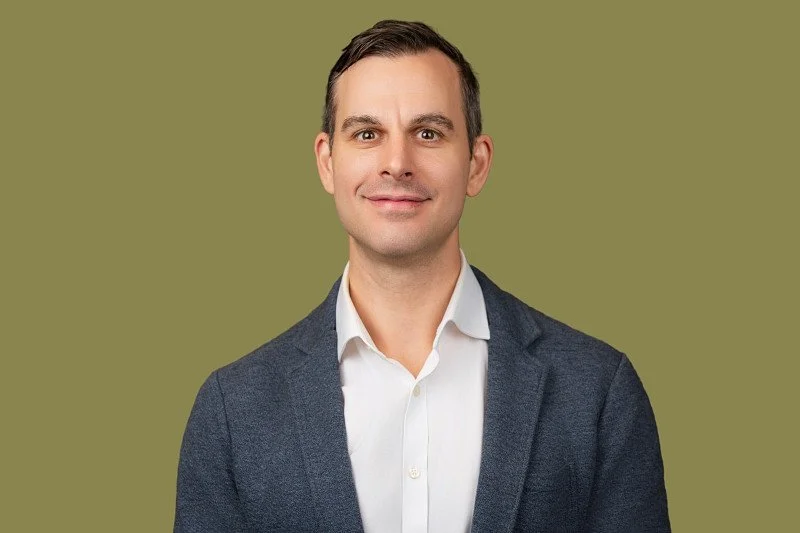 A professional headshot featuring a man in a textured blue blazer and white shirt against a muted olive-green background. 