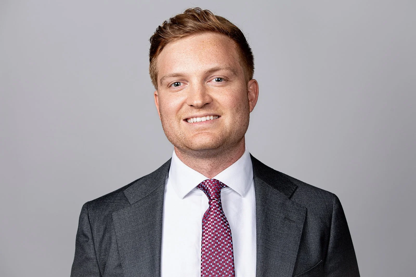A young man with light skin, short brown hair, and blue eyes, wearing a dark gray suit, white shirt, and red patterned tie, smiling against a plain gray background.