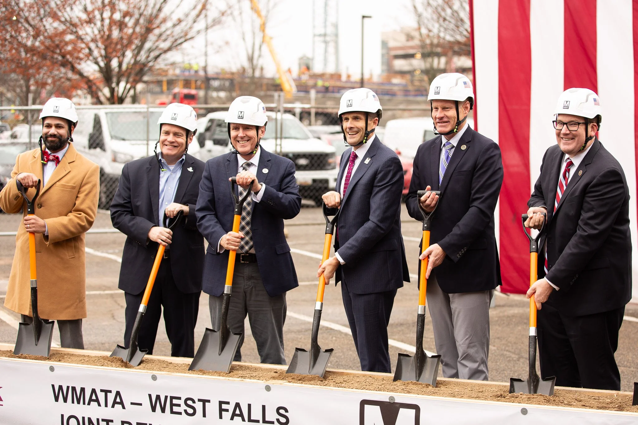 A group of civic and business leaders participate in a ceremonial groundbreaking event for WMATA’s West Falls project, posing with shovels at a construction site. Wearing hard hats and professional attire, the participants celebrate a milestone momen