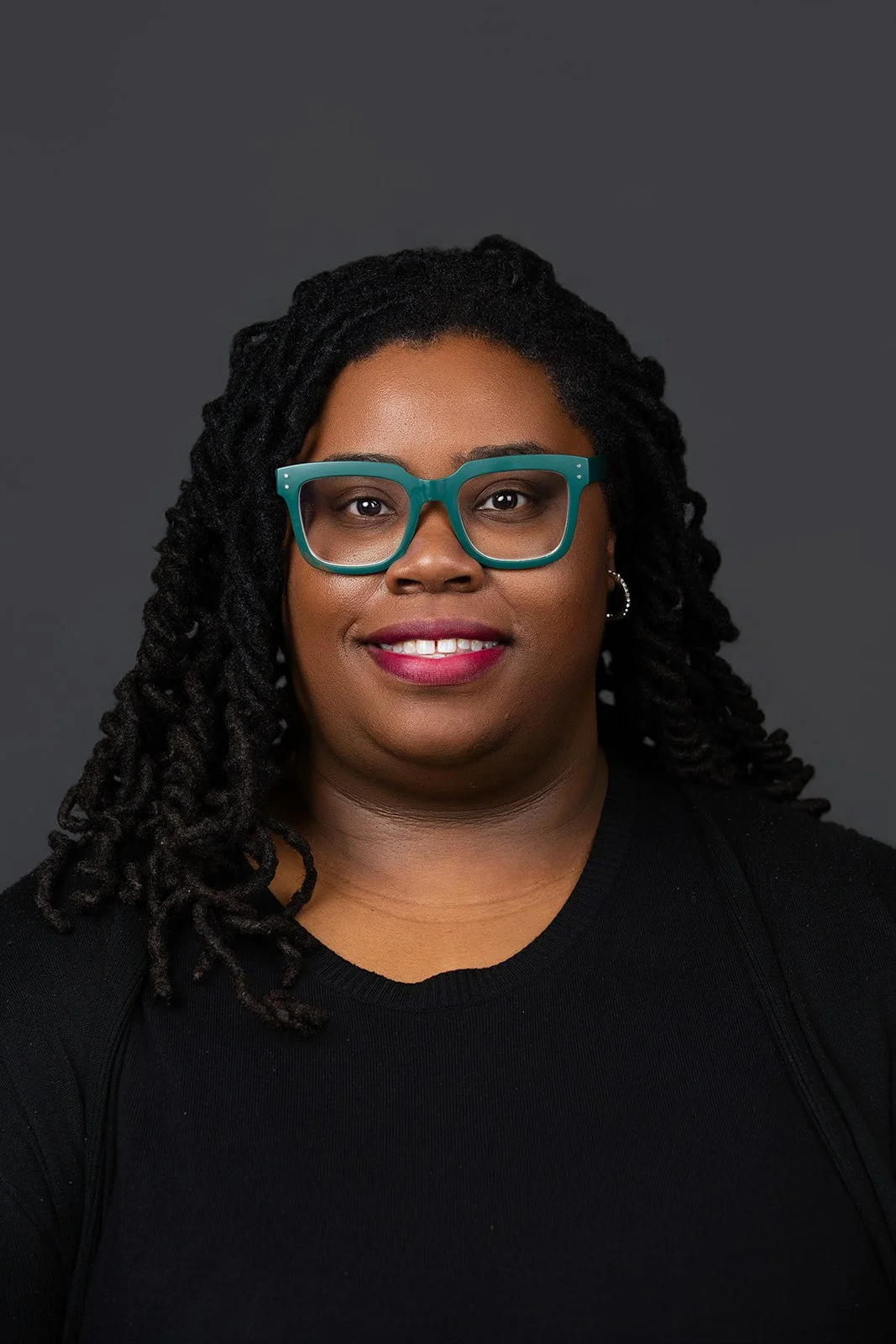 A woman with black twist braids, teal glasses, and a black top, smiling against a dark gray background.