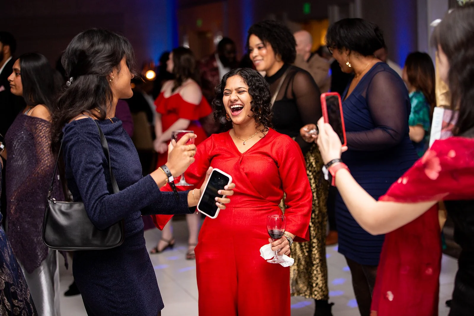 A group of women at a party, with one woman in a red outfit laughing and holding a glass of wine, while others are taking photos and chatting.