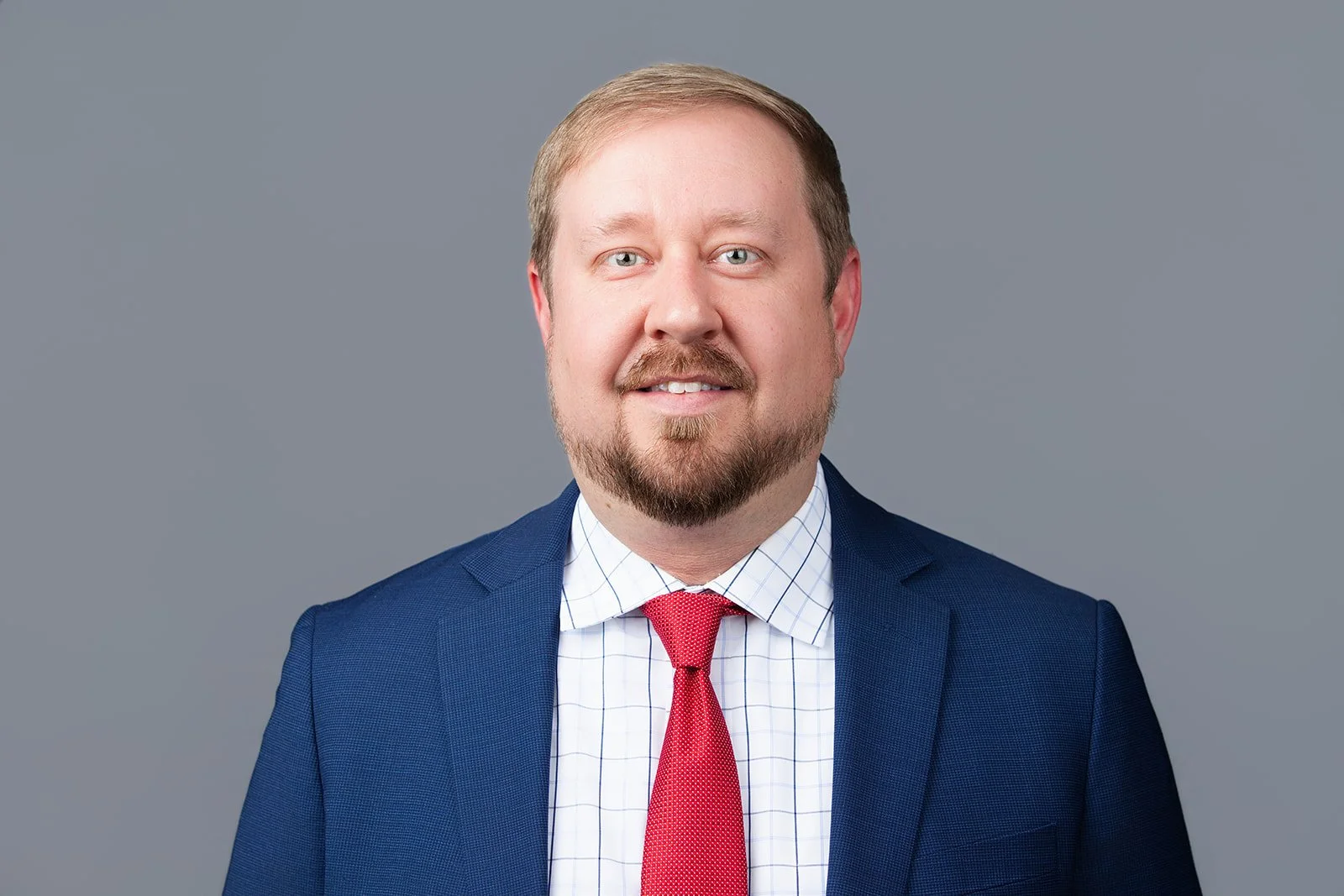 Portrait of a Caucasian man with light brown hair, goatee, wearing a blue suit, white checkered shirt, and red tie, against a gray background.