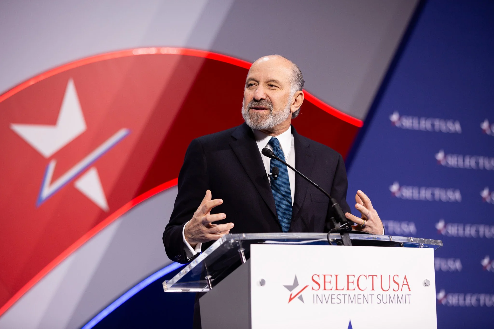 Howard Lutnick delivers remarks at the SelectUSA Investment Summit, addressing attendees from a stage branded with the event’s signature red, white, and blue design. Speaking from a podium, Lutnick shares insights with an audience in a professional c