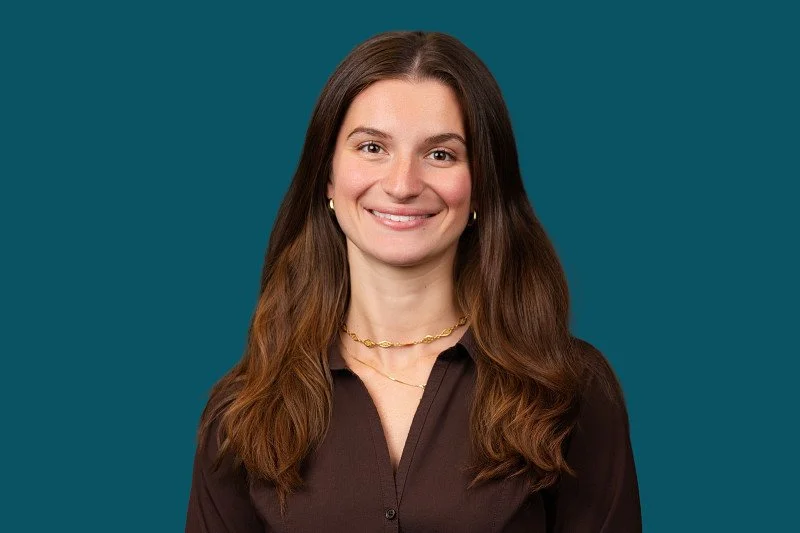 A professional headshot of a smiling woman with long brown hair wearing a dark brown blouse and gold jewelry, photographed against a teal background.
