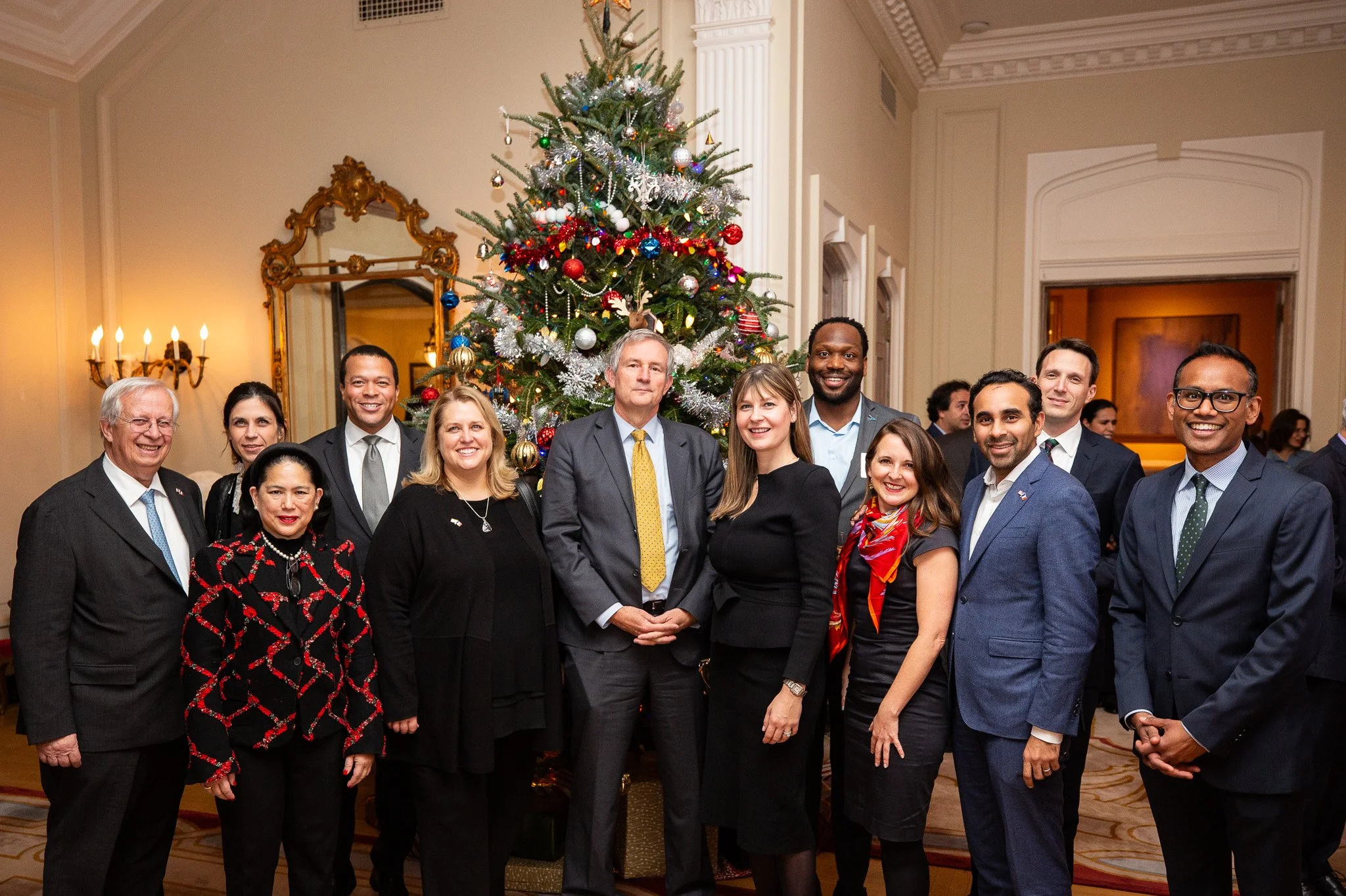 A group of professionals gather for a formal holiday reception in an elegant event space, posing together in front of a decorated Christmas tree. The image captures a moment of connection and celebration among colleagues and community leaders, highli