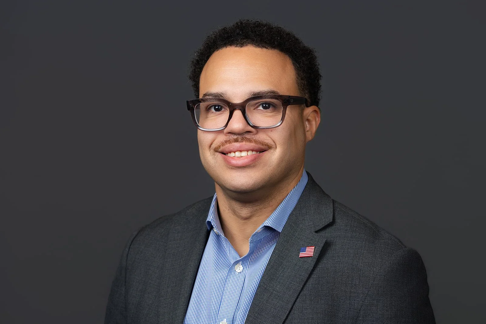 Portrait of a smiling man wearing glasses, a blue collared shirt, and a dark blazer with a small American flag pin on the lapel, against a dark gray background.
