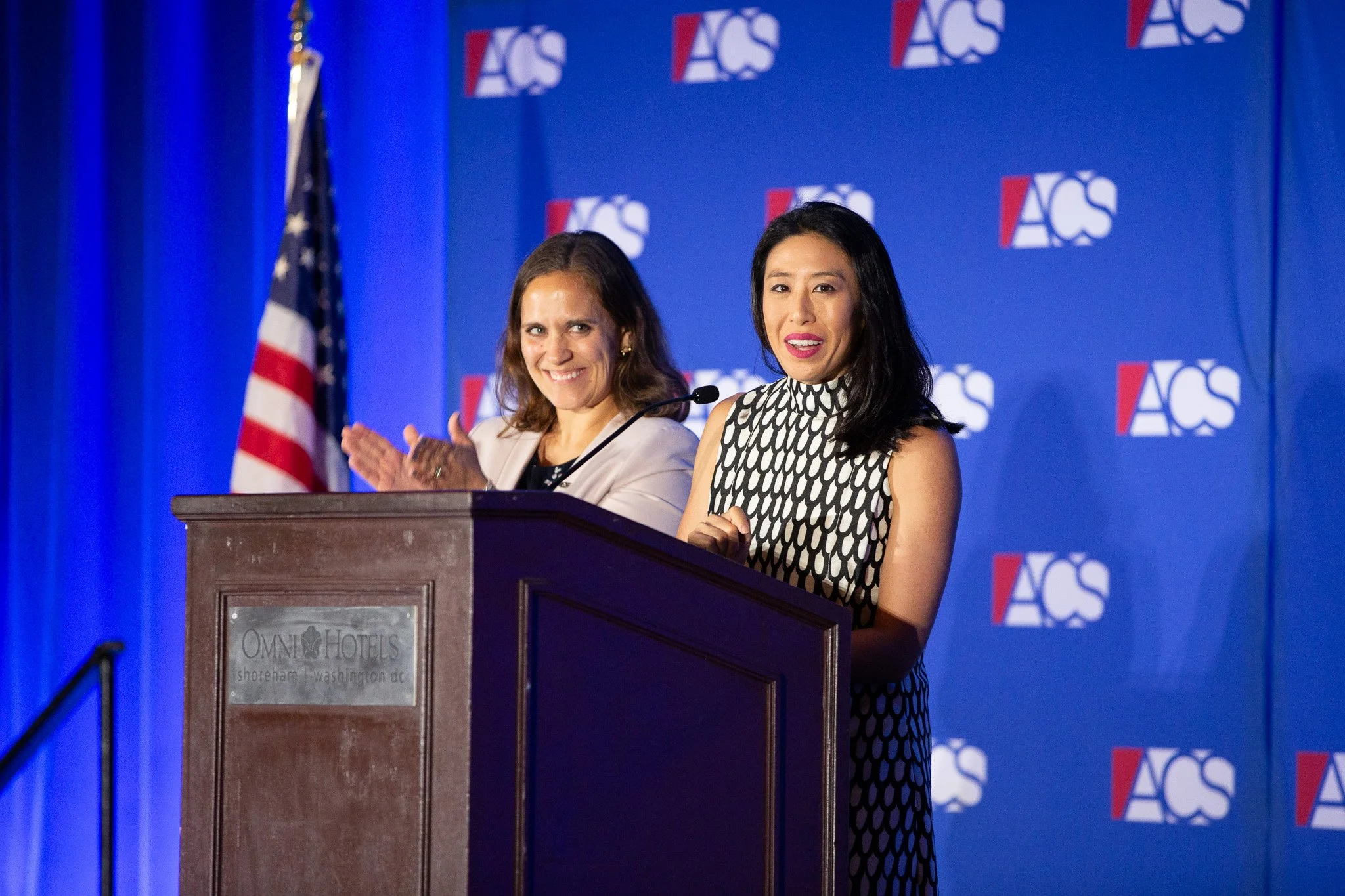 A speaker addresses attendees from the podium during a conference while another presenter applauds beside her. Set against the organization's branded backdrop, the image captures a moment of recognition, leadership, and engagement at a professional i