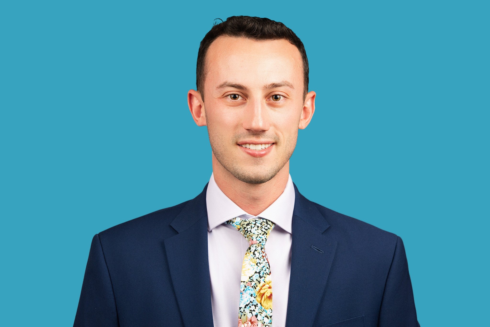 A young man with short dark hair is wearing a bright blue blazer, a white dress shirt, and a colorful floral-patterned tie in shades of blue, yellow, and green, smiling confidently, posed against a teal backdrop. 