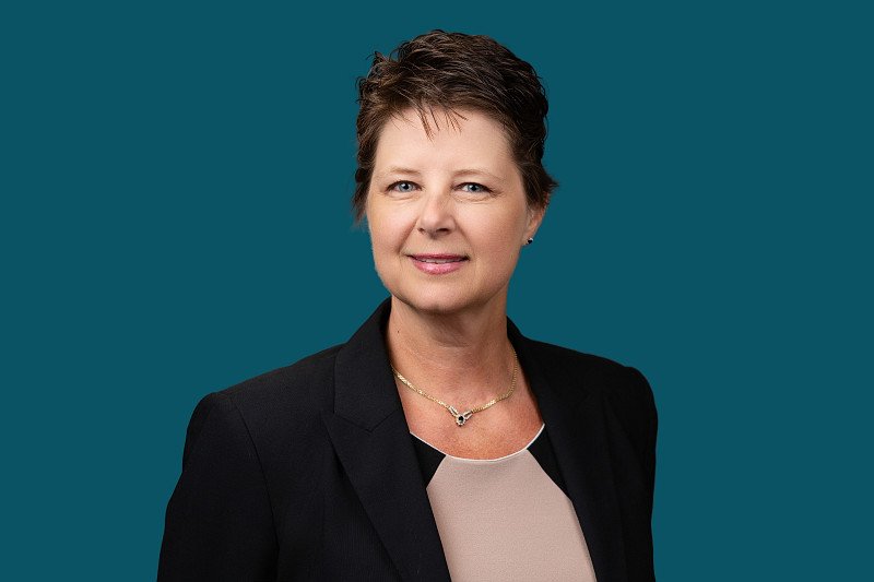 A professional headshot featuring a woman wearing a black blazer and light-colored blouse against a deep teal background. The portrait showcases a warm, approachable expression.