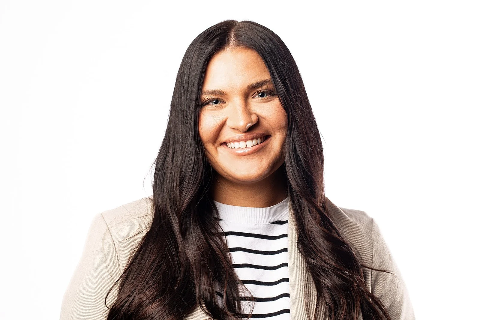 A woman with long dark hair, smiling, wearing a beige blazer and a black and white striped shirt, against a white background.