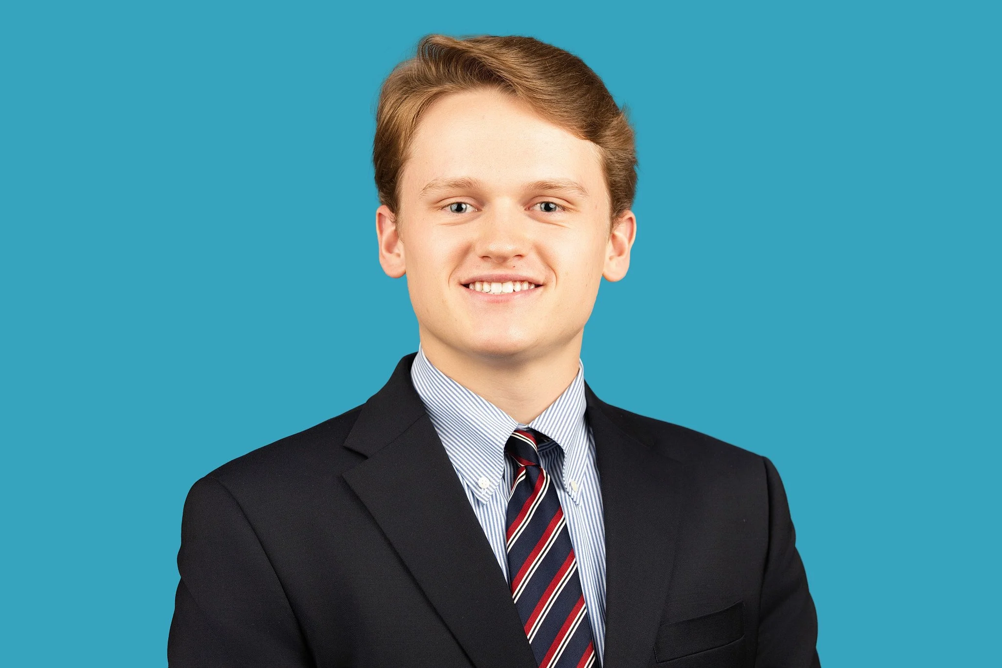 A young man with short light brown hair is wearing a dark navy blazer, a light blue striped dress shirt, and a red, white, and navy striped tie, smiling broadly, posed against a bright turquoise background.