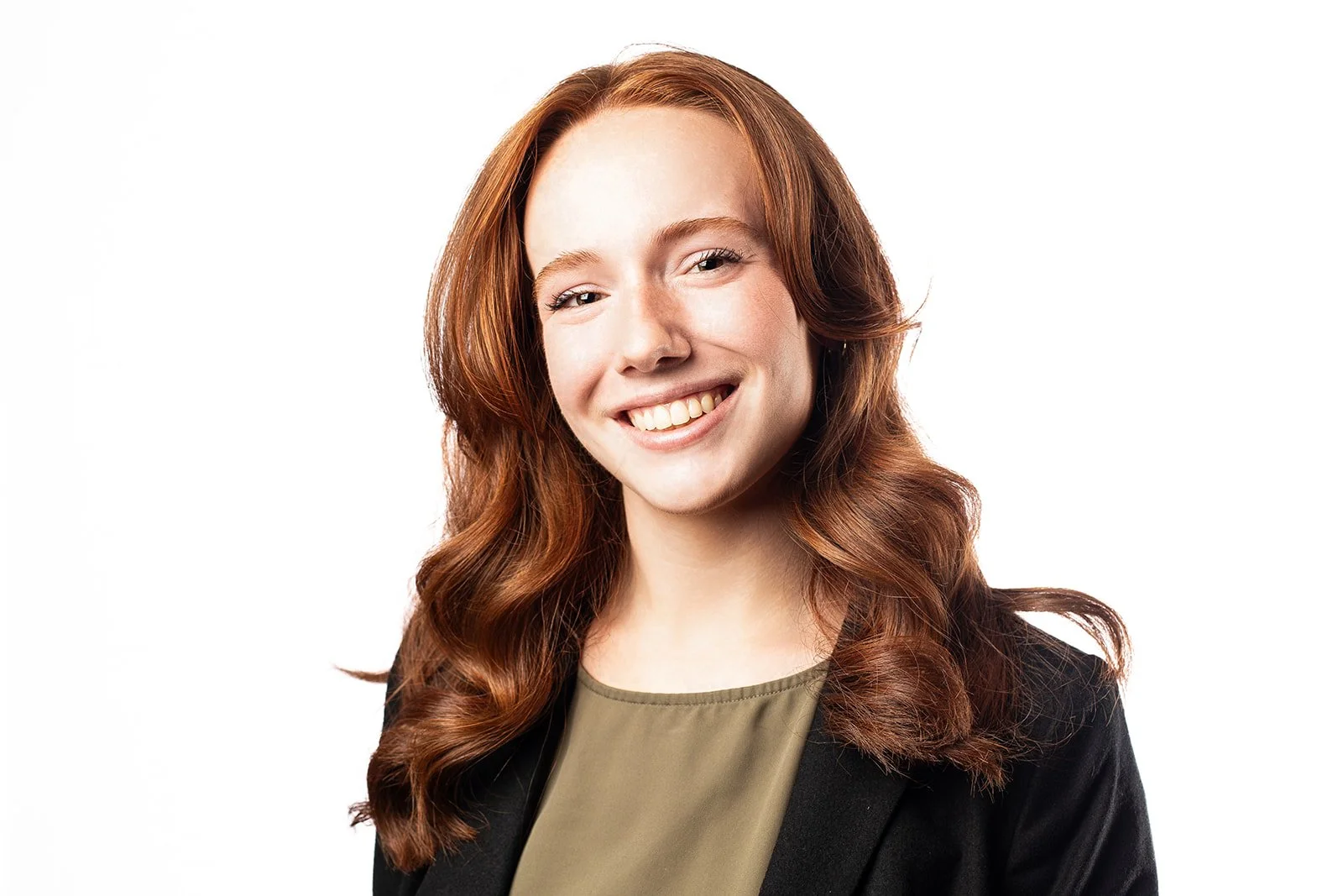 A woman with long wavy red hair smiling, dressed in a black blazer and olive-green top, against a white background.