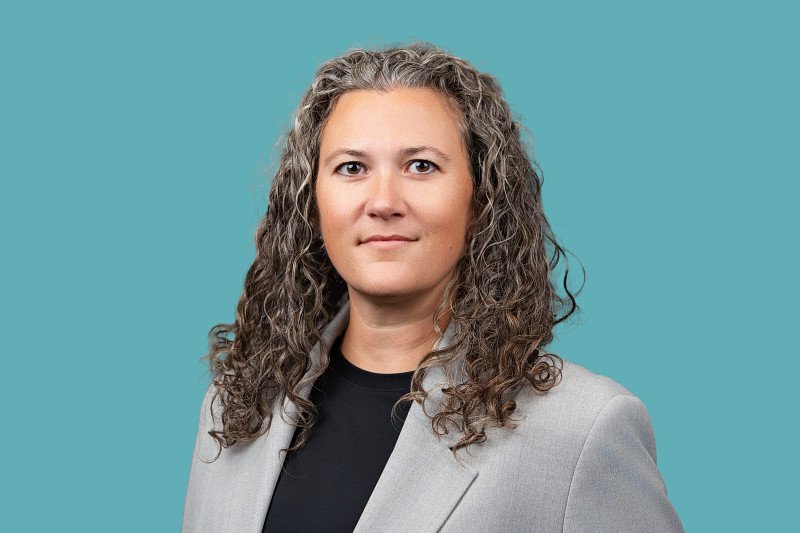 A professional headshot featuring a woman with curly gray hair wearing a light gray blazer and black top against a soft teal background.