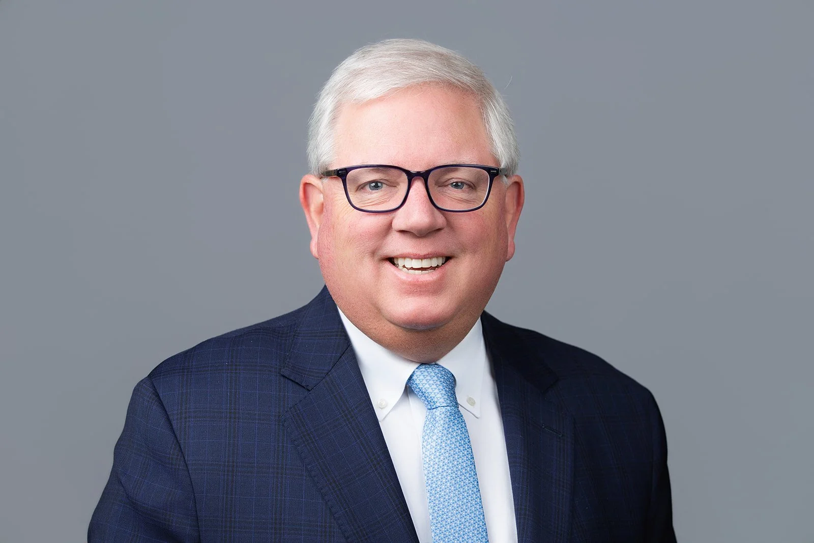 Professional headshot of a smiling middle-aged man with glasses, wearing a navy suit, white shirt, and light blue tie, against a plain gray background.