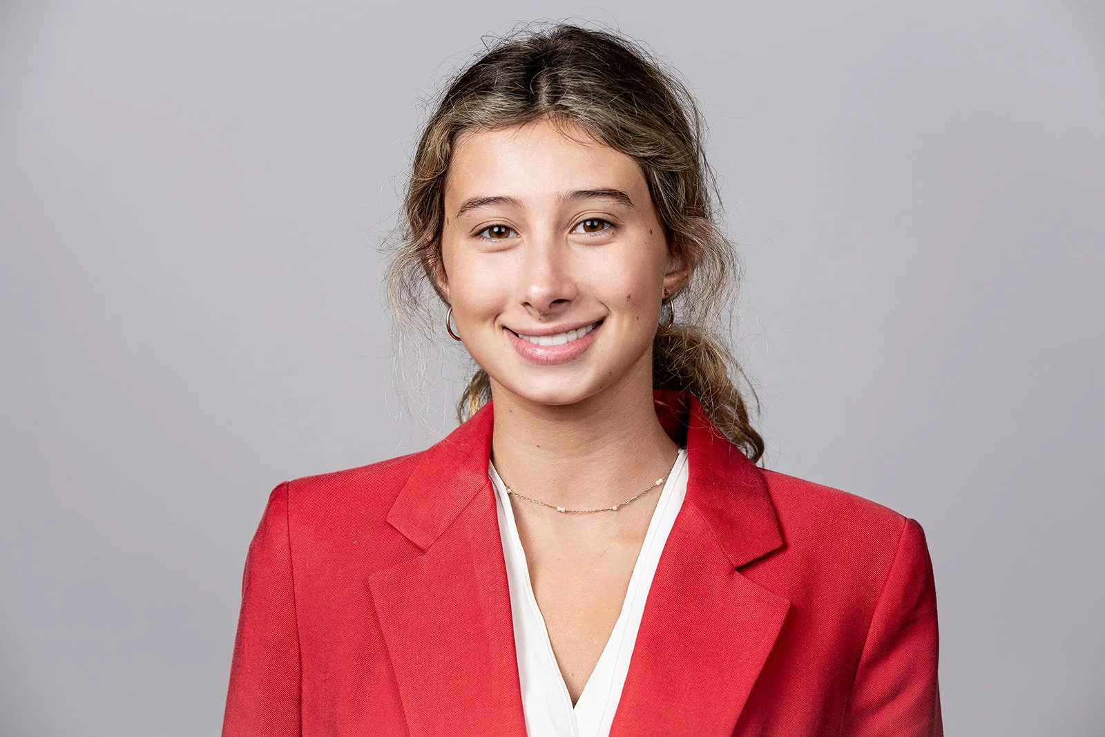 A young woman with light skin, light brown hair tied in a ponytail, wearing a red blazer and a white shirt, smiling against a plain gray background.