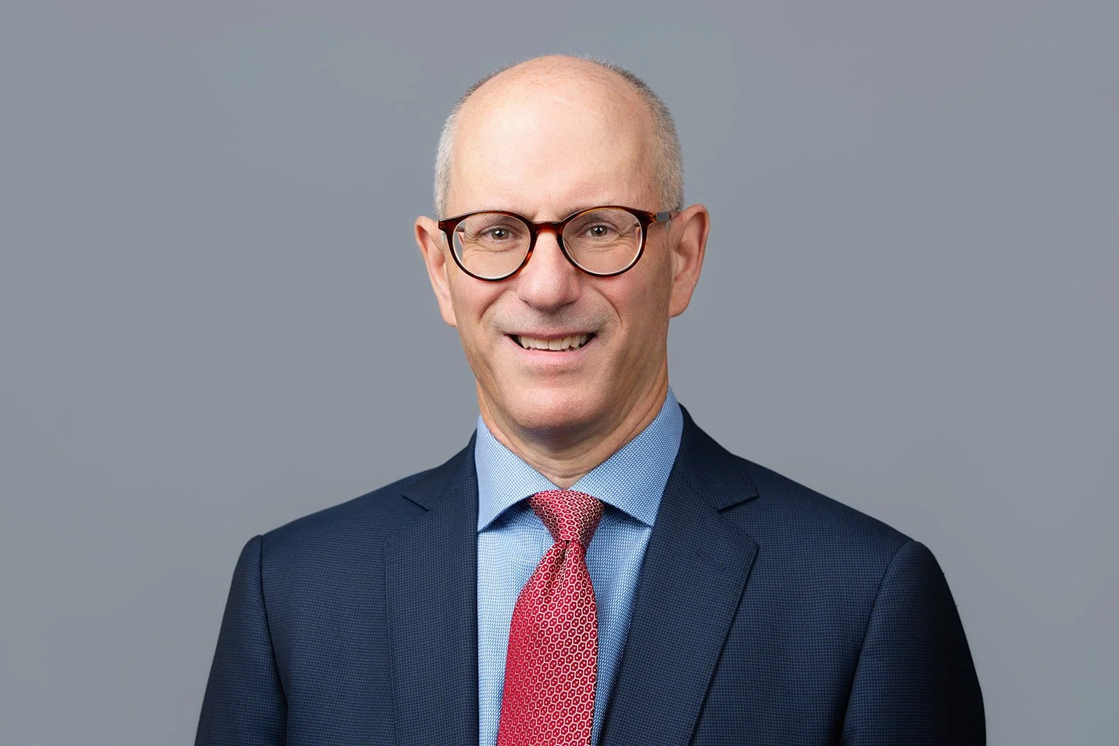Professional headshot of a middle-aged man with glasses, wearing a navy suit, light blue shirt, and red patterned tie, against a gray background.