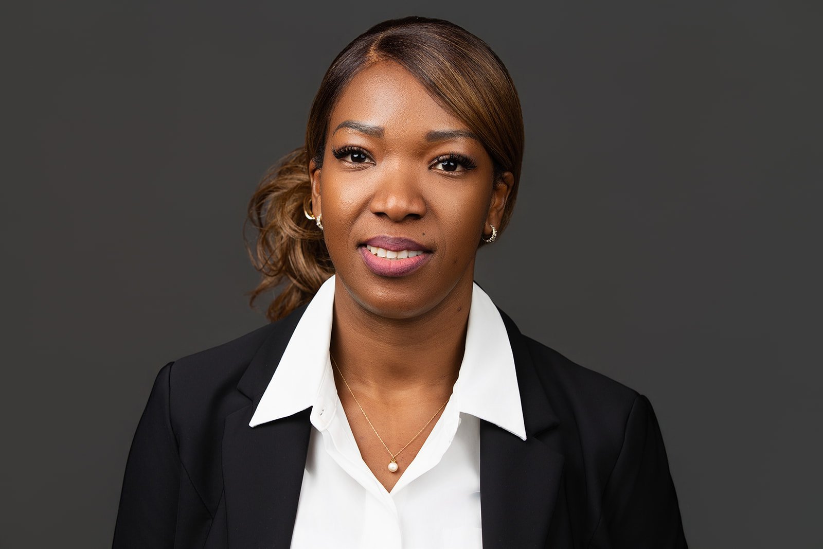 Portrait of a woman with brown hair, wearing a white blouse, black blazer, pearl necklace, and earrings, against a dark gray background.