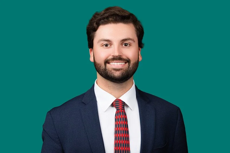A professional headshot of a smiling man with a beard wearing a navy suit, light gray dress shirt, and red patterned tie, photographed against a teal background.