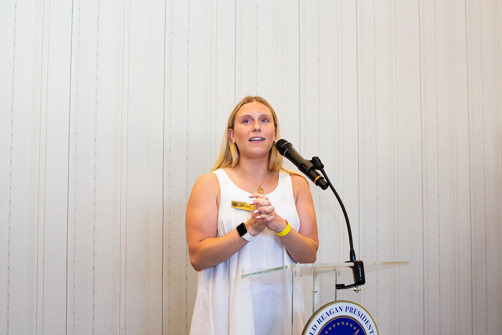 A blonde woman in a white sleeveless top stands at a podium, speaking into a microphone. She appears to be giving a presentation or speech, gesturing with her hands while addressing an audience. Behind her is a white wooden paneled wall. The podium a
