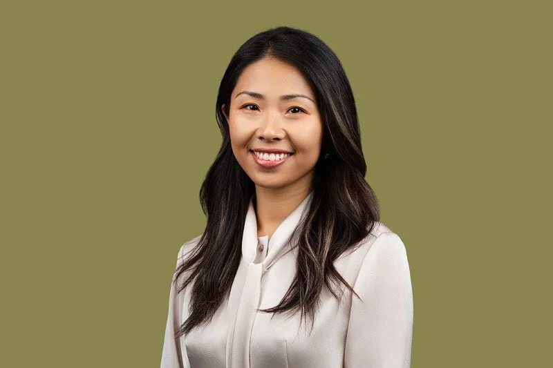 A professional headshot featuring a woman wearing a white blouse against a muted olive-green background. The headshot showcases a  natural smile. 