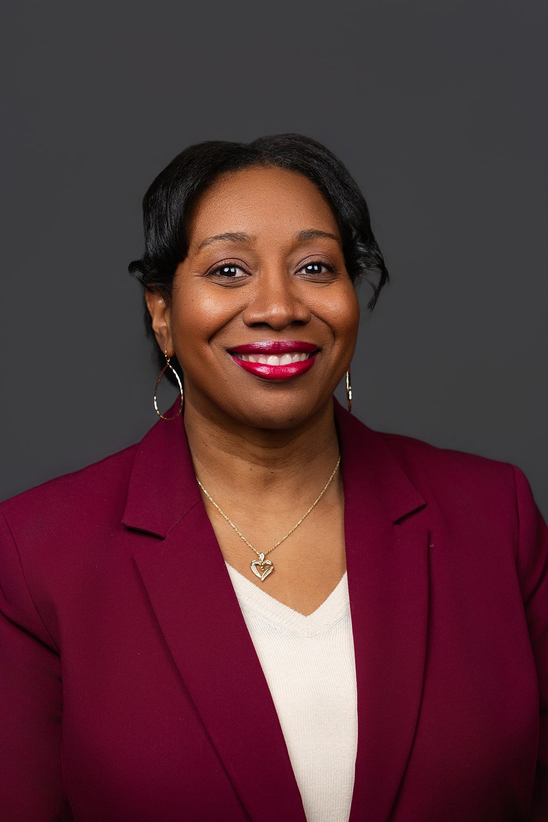 A woman with short black hair, wearing a maroon blazer, white top, gold hoop earrings, and a heart-shaped pendant necklace, smiling against a dark gray background.
