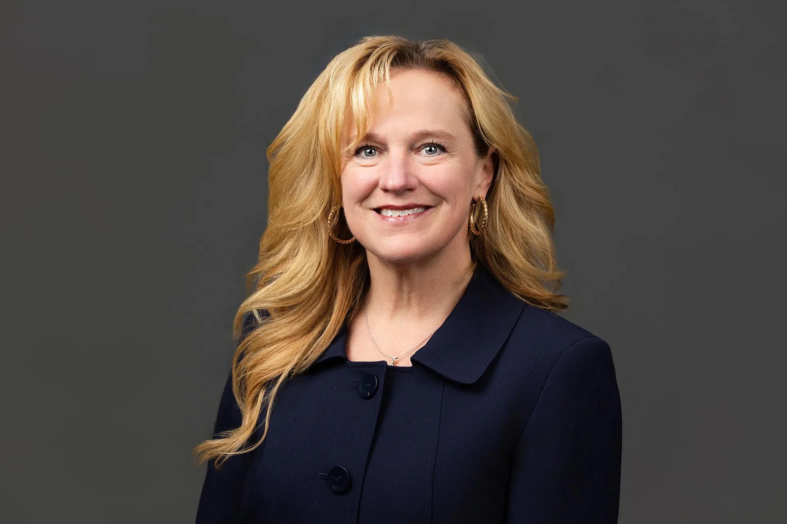 Headshot of a smiling woman with wavy strawberry-blonde hair, wearing a navy blazer and standing against a gray background.