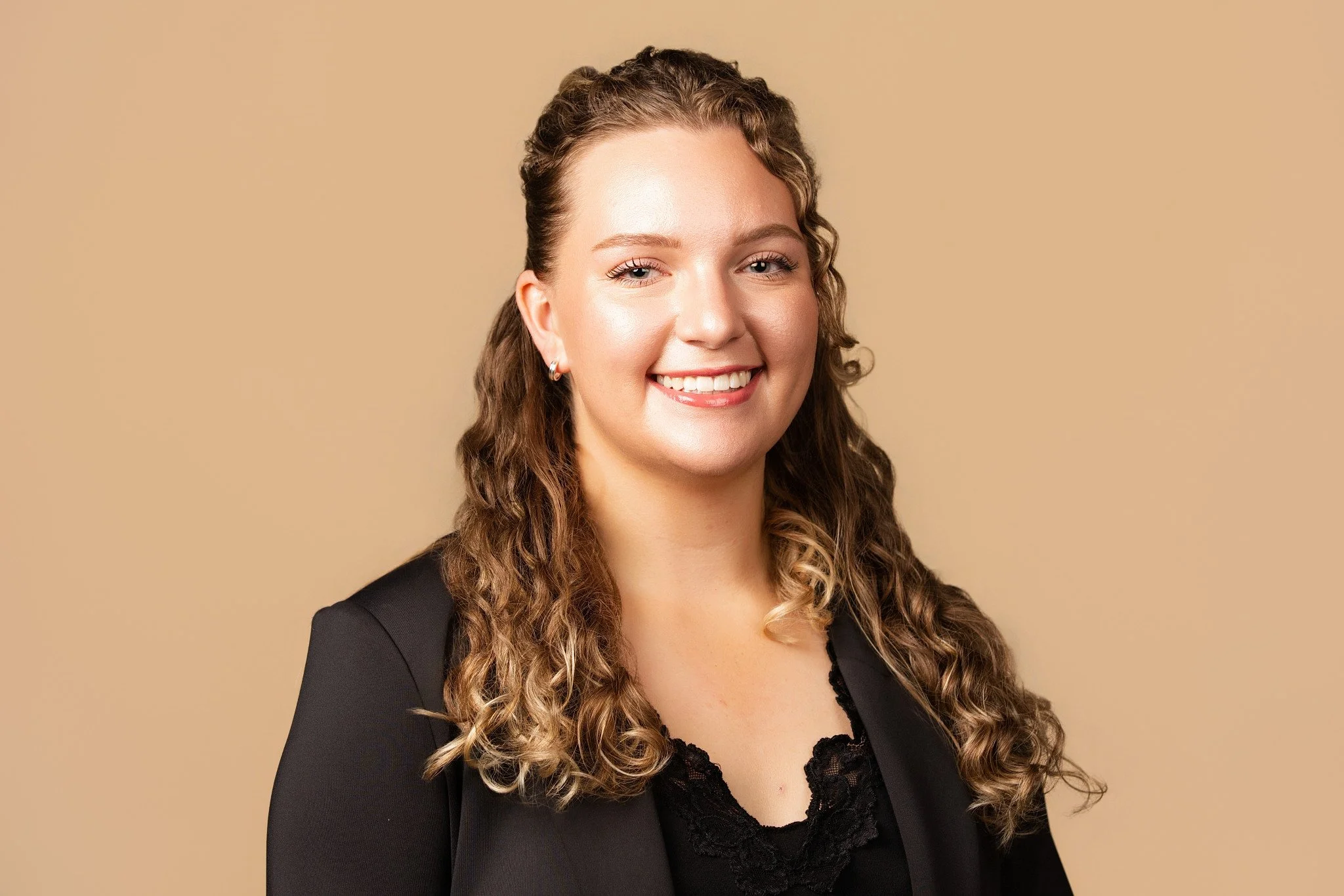 A woman with curly brown hair smiling against a beige background, wearing a black blazer and lace top.