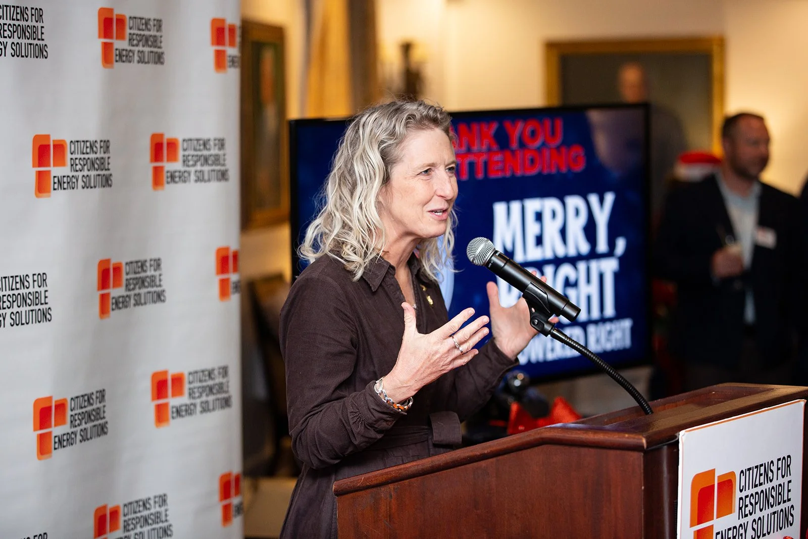 A woman with blonde hair speaking at a podium during a Citizens for Responsible Energy Solutions event. She is gesturing with her hands, wearing a dark outfit, and standing in front of a backdrop with the organization's logo. In the background, a lar