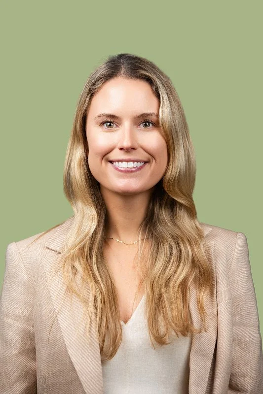 A professional headshot of a smiling woman with long blonde hair wearing a light beige blazer over a cream blouse, photographed against a soft sage green background.