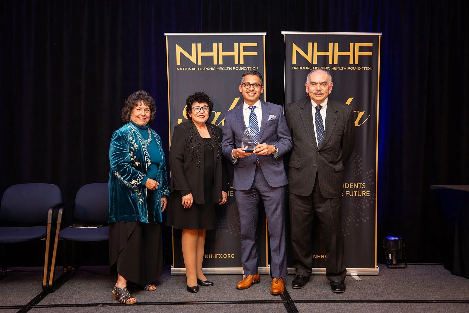 Four people standing on stage at an awards ceremony, with two NHHF banners in the background. The man in the center holds an award.