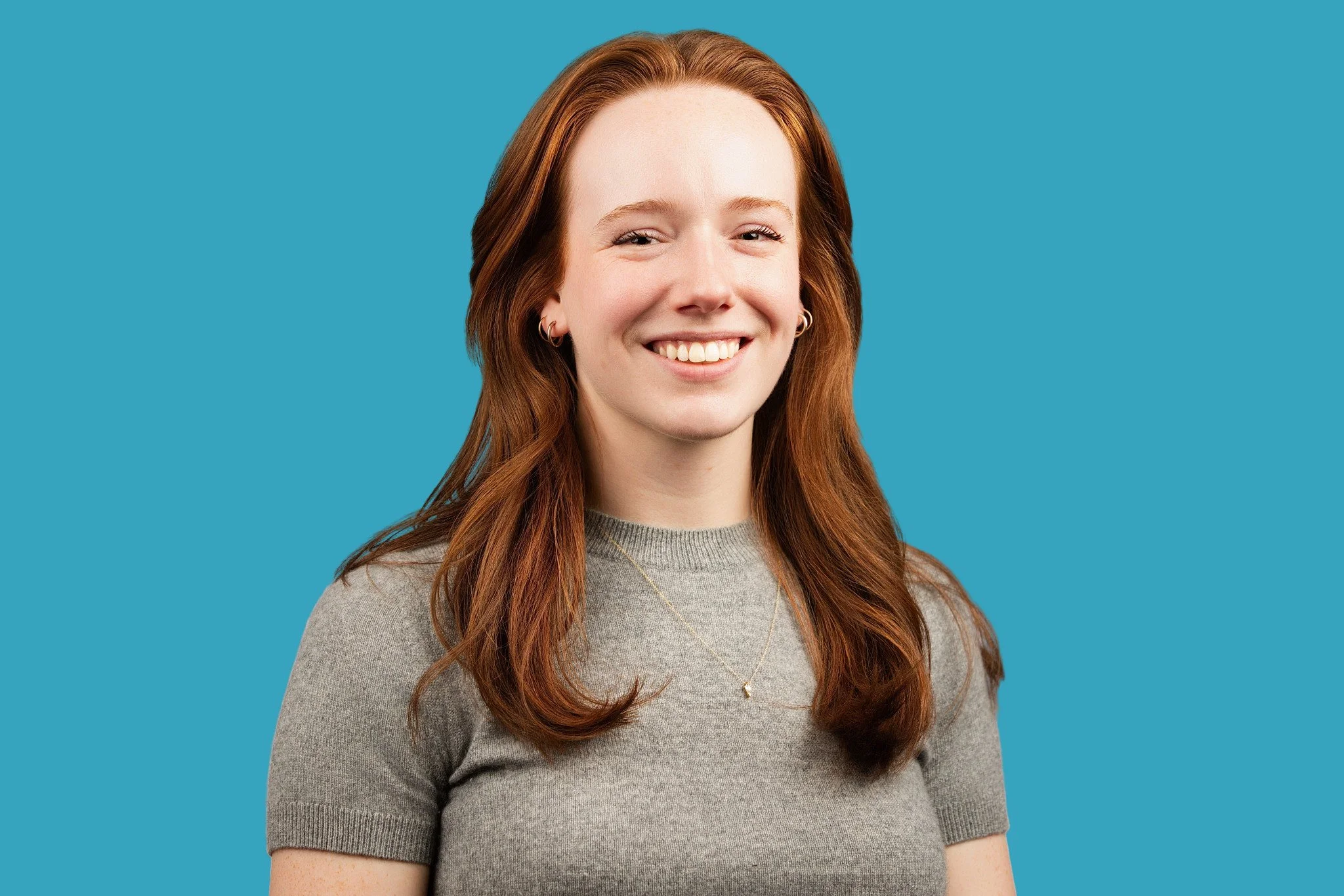 A young woman with long, wavy auburn hair is wearing a grey short-sleeved knit top and a delicate gold necklace, smiling broadly and warmly, posed against a turquoise backdrop. 