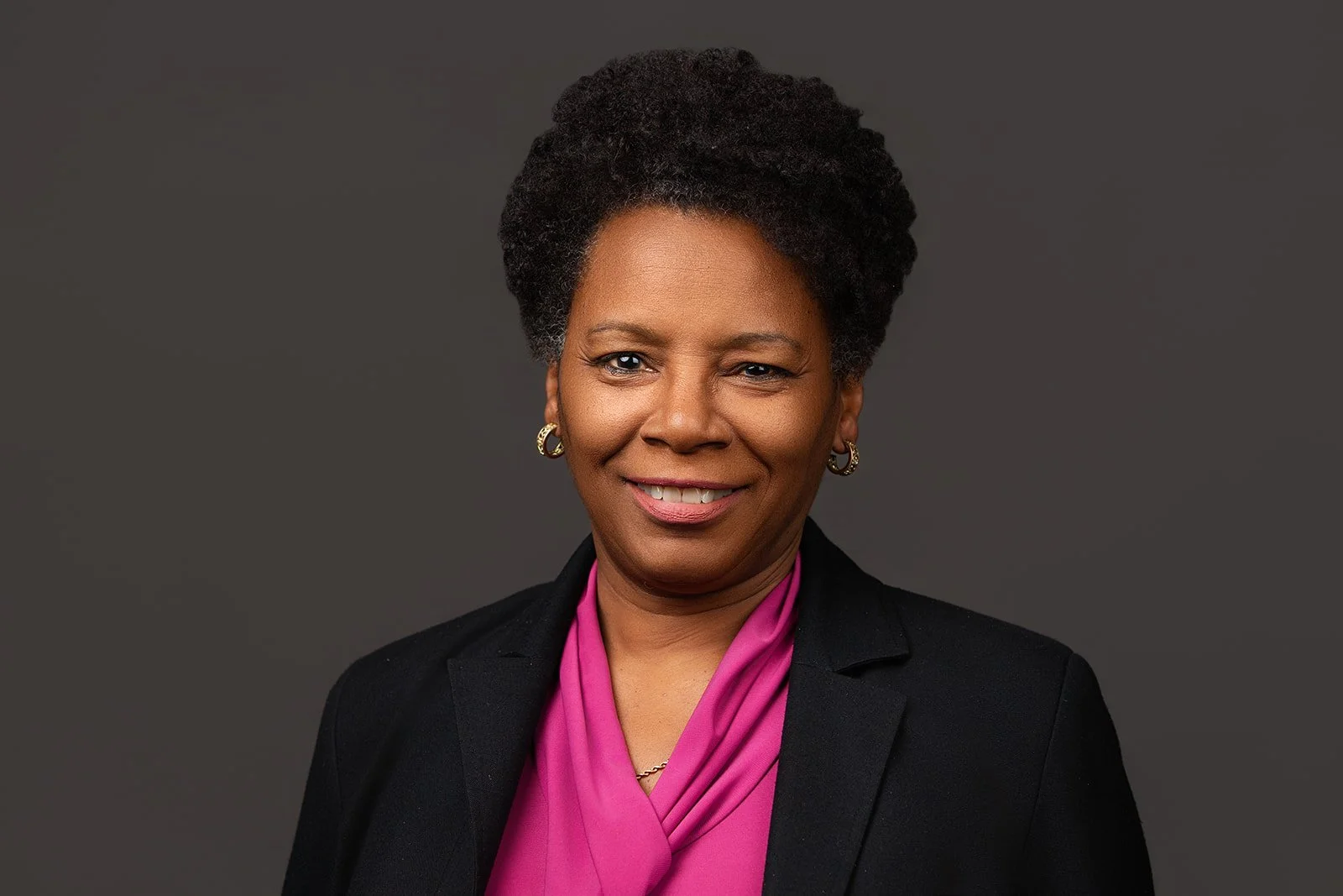 A professional woman with short curly hair smiling at the camera against a dark gray background, wearing a black blazer and a pink blouse.