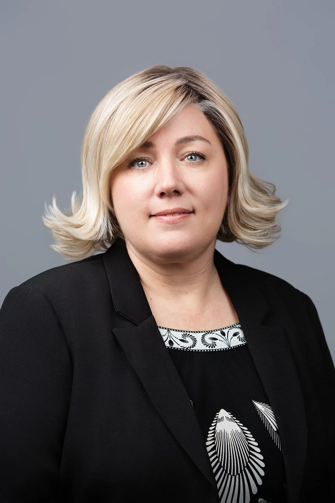 A woman with blonde hair styled in loose waves, wearing a black blazer over a patterned top, against a gray background.
