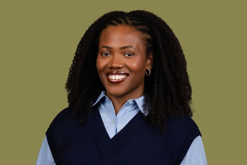 A professional headshot featuring a woman with natural curly hair wearing a navy sweater vest layered over a blue collared shirt against a muted olive-green background. 