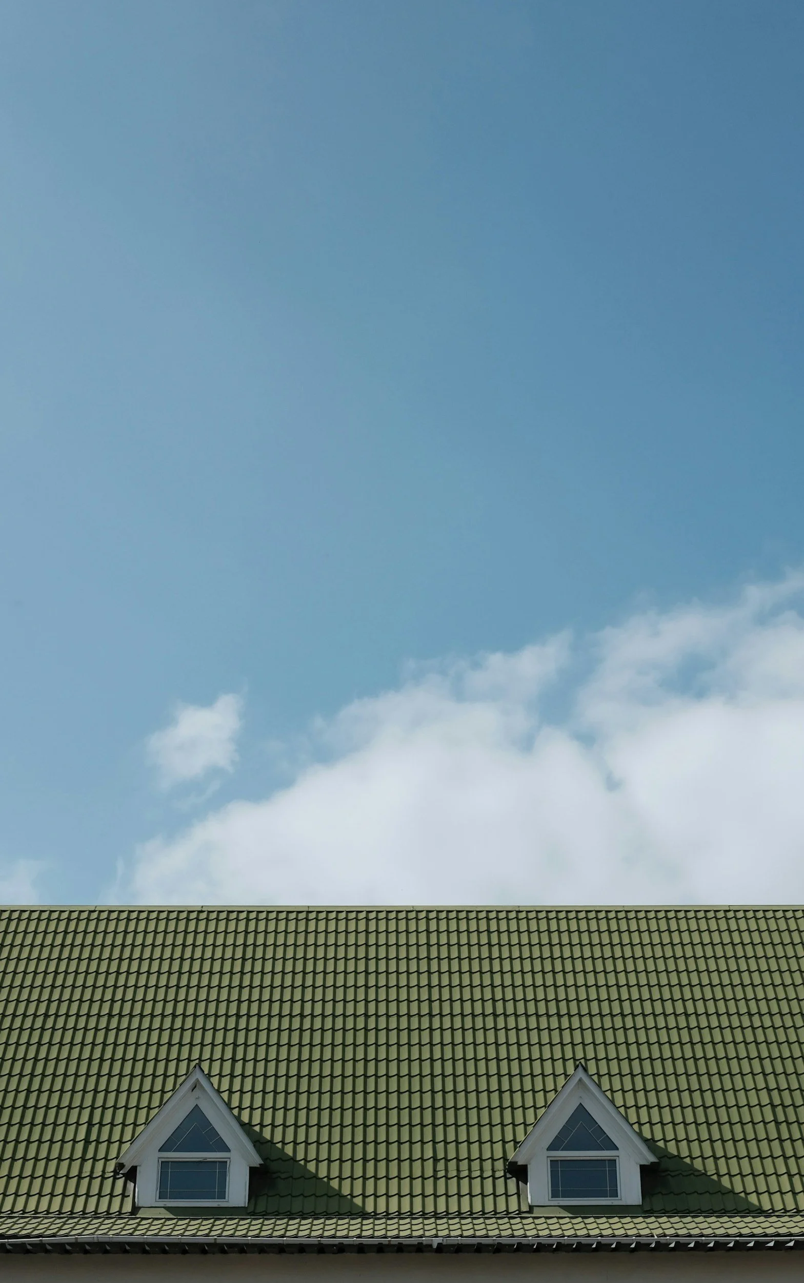 View of a house roof with green tiles and two dormer windows, against a blue sky with some clouds.